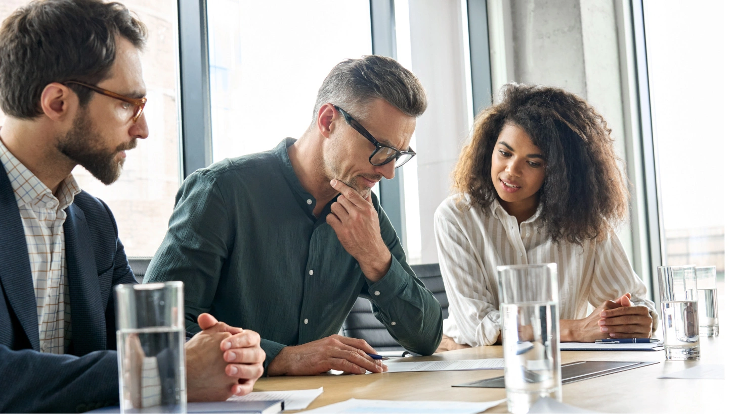 Drei Berufstätige bei einem Meeting im modernen Büro, konzentriert auf Dokumente und Diskussionen, mit transparenten Glastüren im Hintergrund, symbolisiert professionelle Zusammenarbeit.
