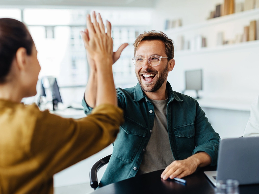 Hochwertige berufliche Interaktion, freundliches High-Five zwischen Kollegen, moderne Büroumgebung, Teamgeist, Zusammenarbeit, positive Arbeitsatmosphäre, beruflicher Erfolg, Teamwork, Kommunikation, moderne Arbeitswelt.