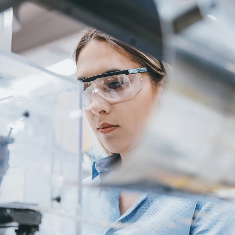 Detaillierte Beschreibung von einer Wissenschaftlerin in einem Labor mit Schutzbrille, die an einer Forschungsarbeit beteiligt ist. Hochwertiges Bild, das Innovation und wissenschaftliche Exzellenz bei ZAG widerspiegelt.