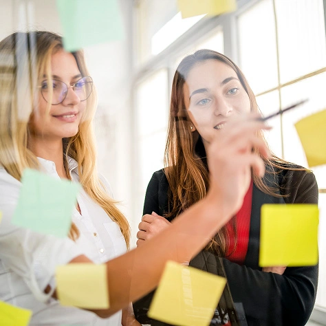 Zwei junge Geschäftsfrauen diskutieren Strategien am Arbeitsplatz mit bunten Haftnotizen.