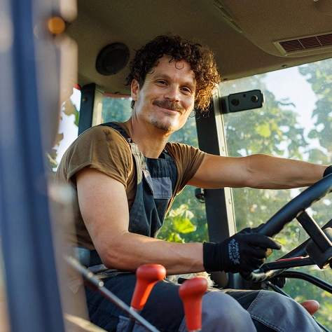 Ein fröhlicher Landwirt fährt in einem Traktor, trägt Arbeitshandschuhe und freut sich auf den Tag auf dem Feld. Perfekt für Landwirtschaft, Maschinen und Natur-Themen.