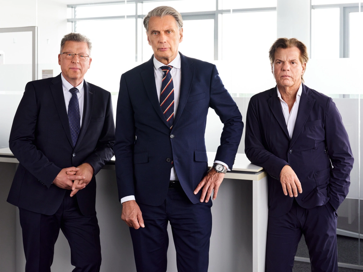 Drei Geschäftsleute in Anzügen stehen in einem modernen Büro. Das Bild zeigt Professionalität, Teamarbeit und Erfolg. Perfekt für Business, Karriere und Unternehmensbranding.