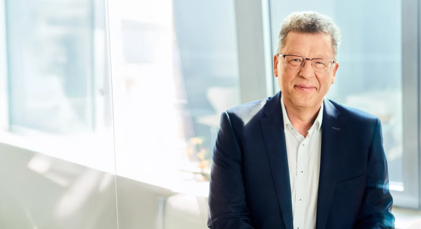 Hochwertiges Business Portrait eines kompetenten Mannes in dunklem Anzug mit Brille, professionell und sympathisch, im modernen Büro mit großem Fenster im Hintergrund.
