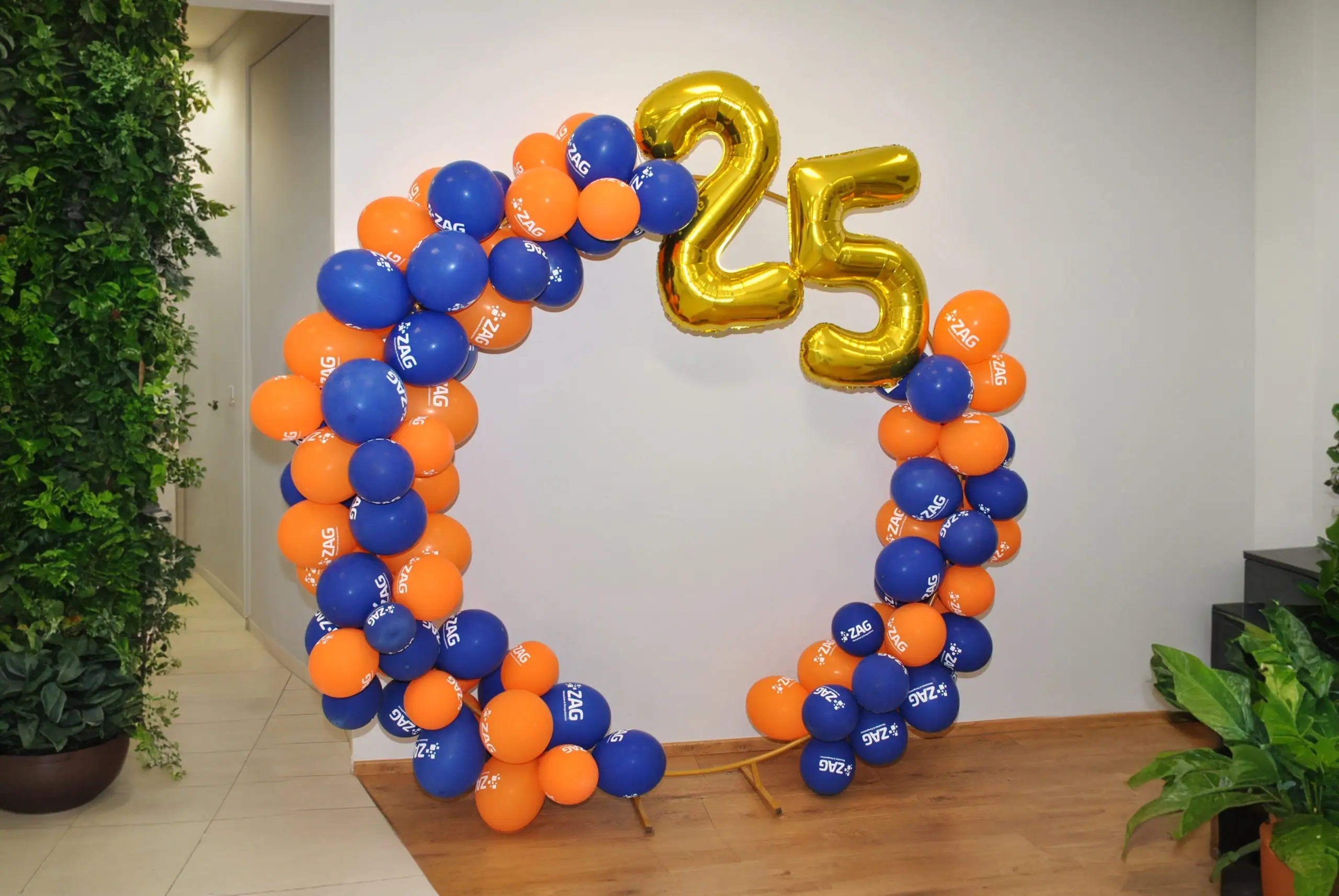 Ballonbogen mit blauen und orangefarbenen Luftballons, goldene Zahlenballons "25", ZAG Firmenlogo, für Jubiläumsfeier, Firmenveranstaltung, Unternehmensjubiläum, Feierdekoration, Jubiläumsfeier, Eventdekoration, Firmenfeier, Anniversary, Balloon Arch, Firmenlogo, Eventdeko, Unternehmensgeschichte.