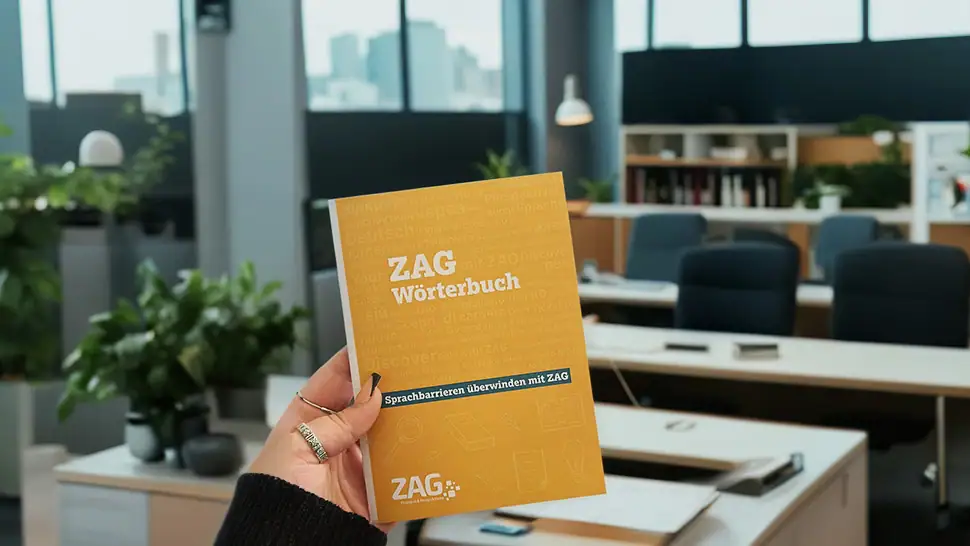 Ein Bild zeigt eine Hand, die ein ZAG Wörterbuch hält, im modernen Büro mit Schreibtischen, Pflanzen und großen Fenstern im Hintergrund, symbolisiert Sprachförderung und Interkulturalität.