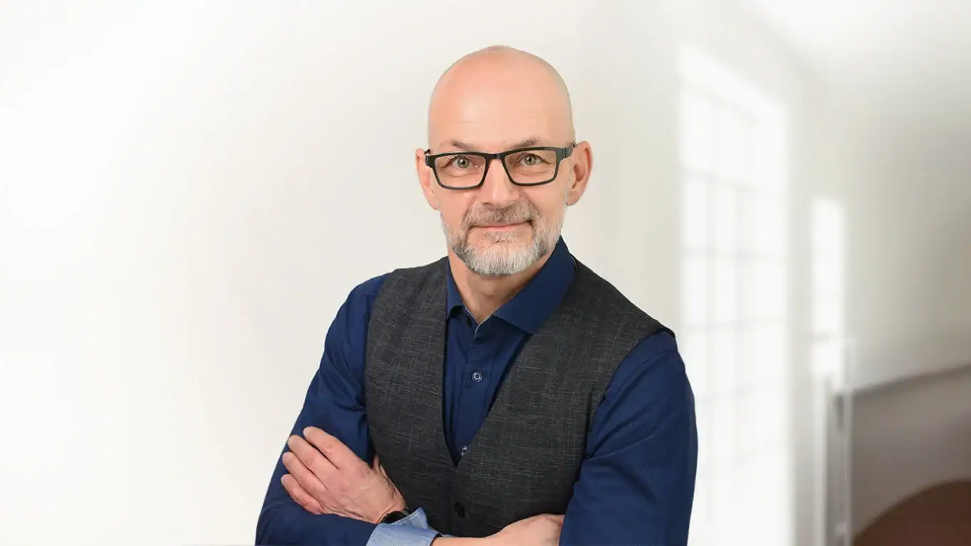 Bewerbungsfoto eines Mannes mit Brille, blauer Hemd und Weste, professionell in hellem Büro setting.