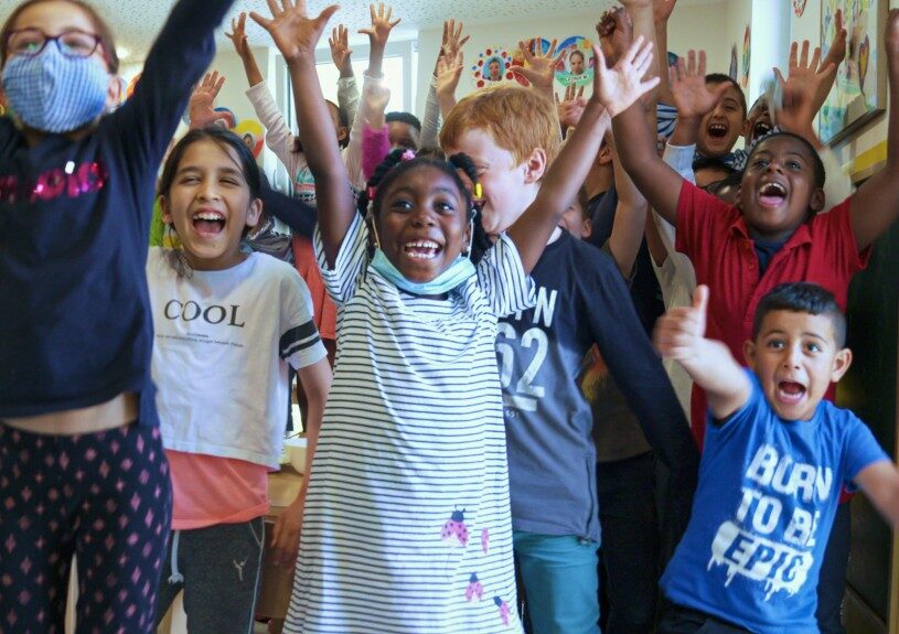 Kinder in einem bunten Klassenraum, die fröhlich lachen und die Arme hochheben, umgeben von kreativen Wanddekorationen, die die Atmosphäre von Gemeinschaft und Lernen bei ZAG widerspiegeln.