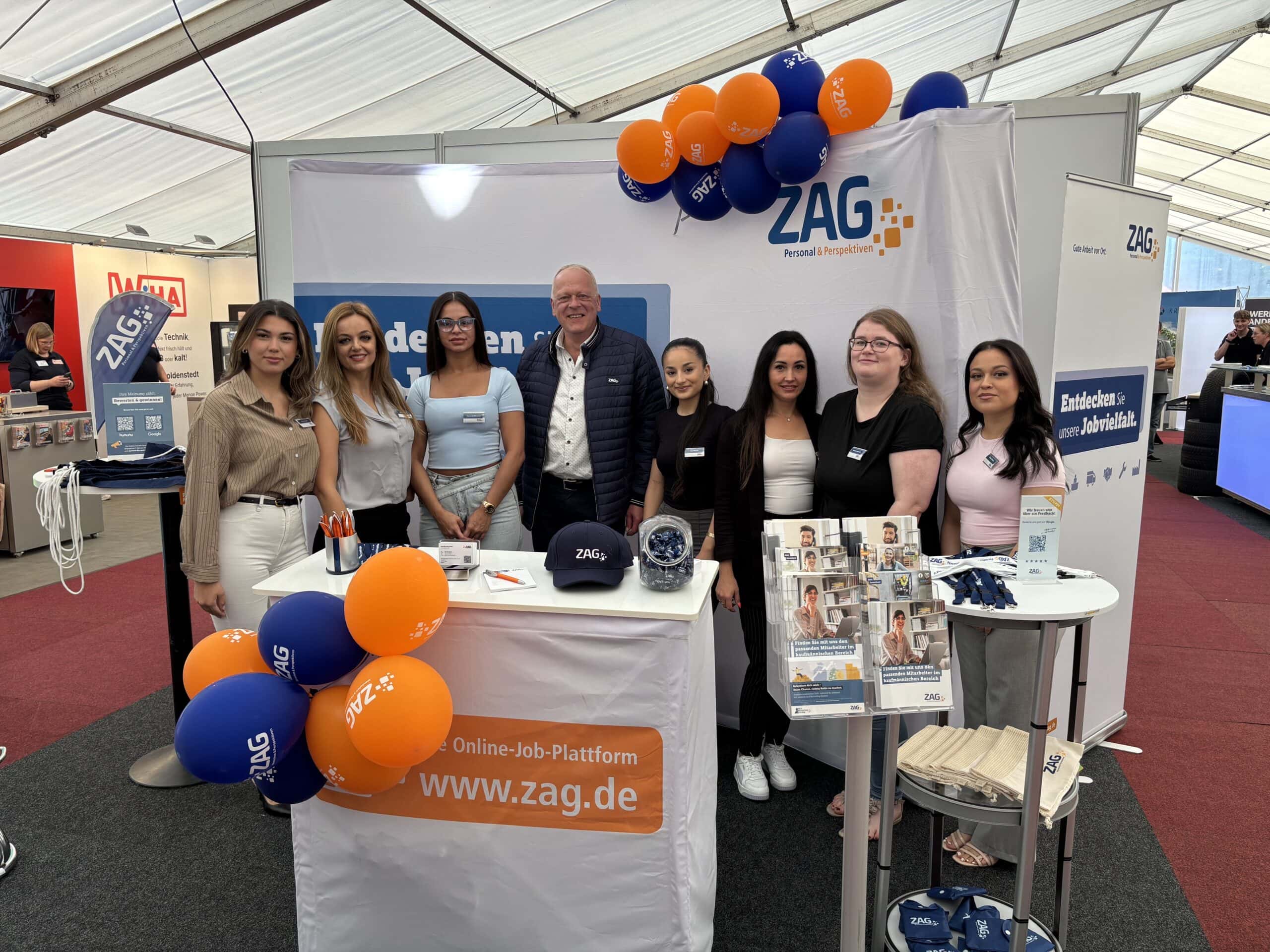 Vielfältiges ZAG-Team bei einer Karrieremesse im Messebau, präsentiert sich an ihrer Messe-Stand mit ZAG-Logo, Flyer-Display und Ballons in den Markenfarben; freundliche Mitarbeiter stehen für Beratung und Jobinformationen bereit.