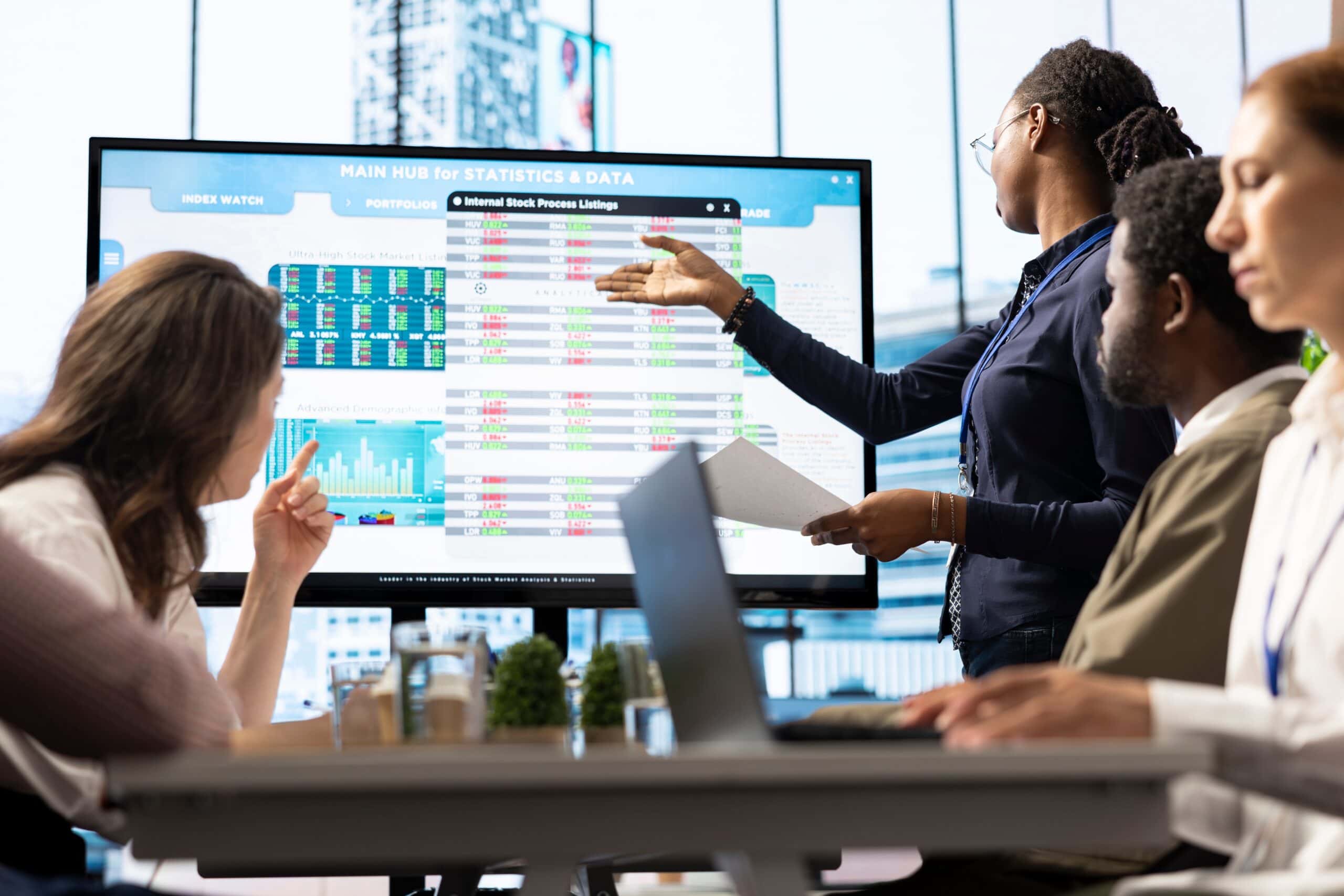 Damen präsentieren einer Gruppe von Kollegen Finanz- und Börsendaten an einem großen Bildschirm in einem modernen Büro.
